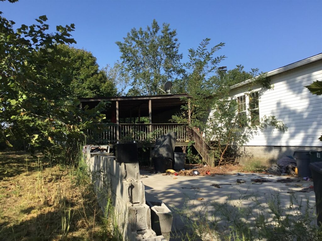 Image of back of manufactured home with large deck and trees growing up in the steps and trash all over patio- our first home