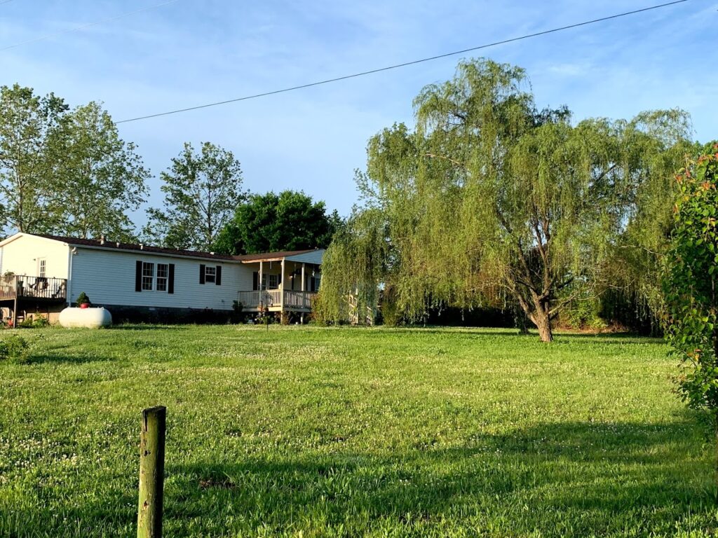 From afar image of white manufactured home with weeping willow in front yard, our first home